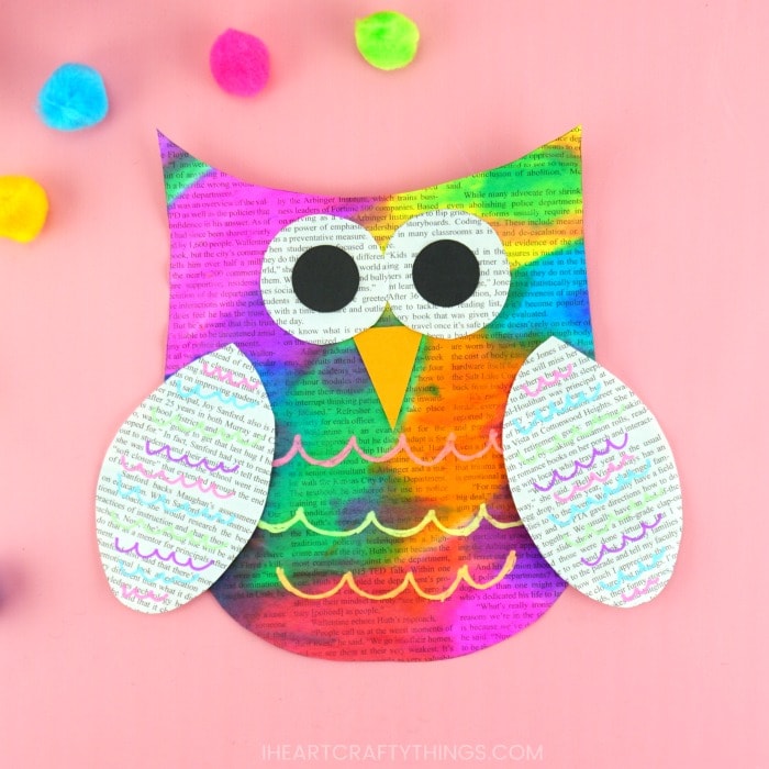 Painted newspaper owl craft laying on a pink background with craft pom poms scattered around.